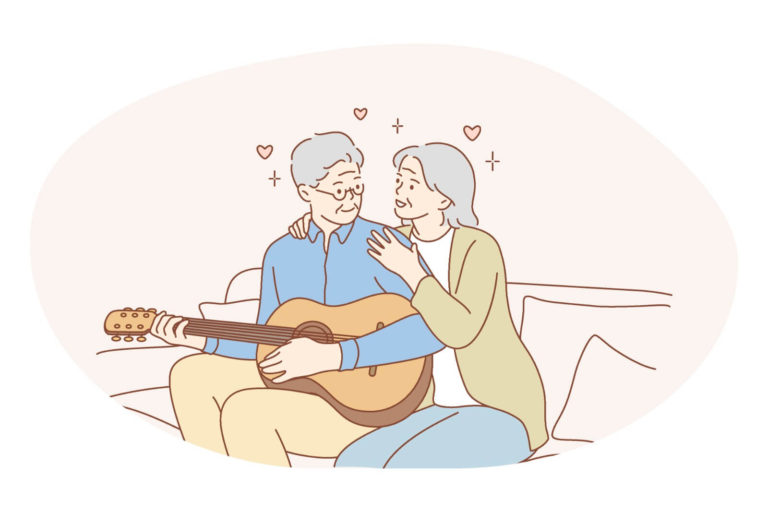 älteres Paar, das auf einem Sofa sitzt, sie umarmt ihn von hinten während er die Gitarre spielt